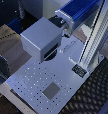 Tête d’un marqueur laser à fibre au-dessus de la table de travail, avec plaque perforée et échantillon métallique.